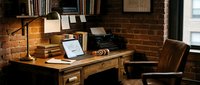 Vintage wooden desk with laptop, desk lamp, and leather chair in brick loft workspace. Creative studio setup for freelancer, designer, or entrepreneur working from home office.