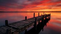 Wooden pier extending into calm water during dramatic sunset with vibrant red and orange sky, long exposure technique creating smooth water reflections for nature and landscape photography.