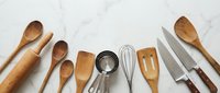 Wooden and metal kitchen utensils arranged on white marble surface including spoons, rolling pin, whisk, spatula and knives for cooking preparation.