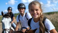 Young female rider smiling during horseback riding lesson outdoors with instructor and fellow student on sunny summer day at equestrian center.