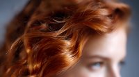 Young woman with flowing auburn hair against dark background, soft focus portrait highlighting natural wavy hairstyle and glowing skin texture.
