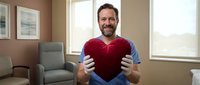 Middle-aged man with beard, Caucasian ethnicity, wearing casual blue shirt, holding red heart shape in living room with warm lighting and comfortable furniture.