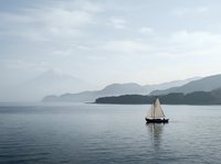Sailboat gliding on calm blue water with misty mountains and iconic volcanic peak in the background on a serene overcast day.
