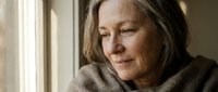 Middle-aged woman with long dark hair and fair complexion, wearing a neutral-toned sweater, gazing thoughtfully out a window with natural light. Suitable for editorial, wellness, and lifestyle content.