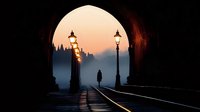 Silhouette of person walking through stone archway at sunset with vintage street lamps creating atmospheric tunnel perspective and warm golden light.