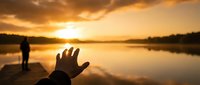 Hand reaching toward golden sunset over calm lake with silhouetted figure in background creating peaceful spiritual moment for meditation and wellness content.