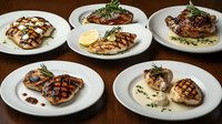 Grilled chicken breasts with various sauces and garnishes on white plates arranged on dark wooden surface, restaurant quality presentation with herbs.