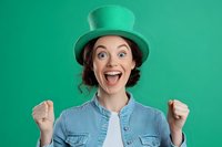 Excited young Caucasian woman wearing green leprechaun hat and denim jacket showing victory gesture against green background, celebrating St Patrick's Day.