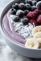 Smoothie bowl with fresh blueberries, raspberries, banana slices and coconut flakes in white ceramic bowl. Purple acai base topped with frozen berries.