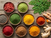 Colorful superfood powders and spices in wooden bowls with fresh moringa leaves, goji berries, ginger and turmeric roots on rustic wood background for healthy cooking.