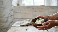 Hands holding wooden bowl with white flower, offering gesture in bright interior space with natural light and rustic wooden table.
