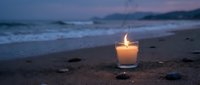 Glowing candle on sandy beach at twilight with ocean waves and purple sky creating peaceful meditation atmosphere for relaxation and mindfulness practices.