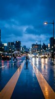 Wet city street at dusk with glowing streetlights and traffic, urban evening scene with reflective pavement and blurred pedestrians crossing road.