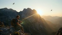 Hiker with backpack sitting on mountain peak at sunrise, watching birds fly over dramatic rocky landscape and valleys below.