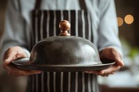 Chef in striped apron holding vintage metal cloche food cover, preparing to reveal gourmet meal in restaurant setting.