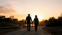 Couple walking with small dog on rural road at golden hour sunset, holding hands while strolling through countryside landscape for lifestyle and relationship content.