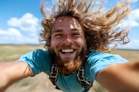 Energetic young bearded man with flying hair taking selfie outdoors on windy day, wearing turquoise t-shirt against summer landscape with blue sky and hills.