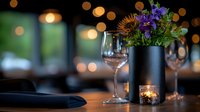Elegant restaurant table setting with black matte vase holding purple wildflowers, wine glass and candle creating romantic ambiance against blurred bokeh lights.