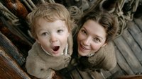 Mother and toddler son wearing matching beige sweaters looking up with surprised expressions while lying on wooden deck with rope details.