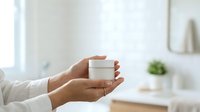 Woman with medium skin tone holding white cosmetic jar in bright bathroom setting, suitable for beauty, skincare, and wellness product marketing.