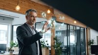 Mature Caucasian businessman in suit touching transparent digital interface with glowing icons in modern office environment with wooden ceiling and pendant lights.