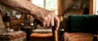 Elderly man's weathered hands gripping wooden walking cane in cozy home interior. Senior mobility aid for independence and stability in daily living.