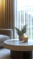 Minimalist living room corner with round coffee table, palm leaves in ceramic pot and glowing candle against sunlit vertical blinds and grey sofa.