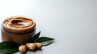 Creamy peanut butter spread in glass jar with fresh green leaves and whole peanuts on gray background, natural food photography styling.