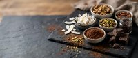 Assorted spices and chocolate ingredients arranged on dark slate board with wooden bowls containing cocoa powder, nuts, and aromatic seasonings for cooking.
