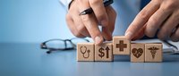 Adult male hands arranging wooden blocks with healthcare symbols including stethoscope, dollar sign, and medical icons on blue desk surface with glasses nearby.