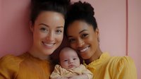 Two happy mixed-race women in yellow sweaters holding newborn baby against pink background. Family portrait with mothers and infant child.