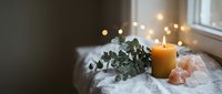 Cozy home spa setup with burning candle, eucalyptus branch and pink salt crystals on marble surface near window with warm bokeh lights creating relaxing atmosphere.