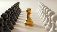 Golden chess pawn stands between rows of black and white pawns on neutral background representing leadership, strategy and standing out from crowd.