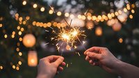 Two hands holding a lit sparkler against a blurred bokeh background with warm golden lights, suitable for celebration, festive events, New Year, and holiday party concepts.