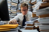 Caucasian businessman in glasses overwhelmed by paperwork stacks at office desk, showing stress and burnout while examining documents. Workplace pressure concept.