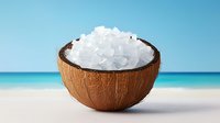 Coconut shell bowl filled with coarse white sea salt crystals on tropical beach with turquoise ocean and clear blue sky background for spa and wellness concepts.