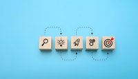 Business strategy concept with wooden blocks showing search, innovation, launch, teamwork and target icons connected by dotted arrows on blue background.