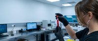 Female scientist of East Asian descent examining a dark laboratory sample or specimen in a gloved hand within a modern research facility equipped with computers and equipment.
