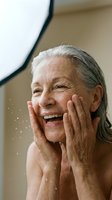 Senior woman with gray hair washing face with water, smiling joyfully while touching cheeks with both hands in bathroom setting for skincare and wellness content.