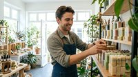 Caucasian male florist or plant shop owner in apron arranging potted plants on wooden shelves in bright, plant-filled retail space with natural light from windows.