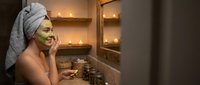 Young woman applying green facial mask in modern bathroom with towel on head enjoying spa skincare routine at home for wellness and beauty treatment.
