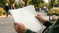 Senior man with weathered hands holding blank white newspaper outdoors in park setting with blurred background, suitable for editorial mockups and lifestyle content.
