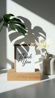 Thank you card in wooden stand with white lily and monstera leaf shadow, minimal elegant decor for gratitude, appreciation, and greeting concepts.