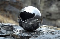 Polished metallic sphere reflecting dramatic rocky landscape against blurred natural background, creating artistic contrast between smooth surface and rugged terrain.