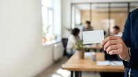 Businessman in dark suit holding blank white business card in modern office environment with colleagues working in background, suitable for corporate branding and professional networking concepts.