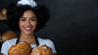 Young African American woman baker in white uniform holding fresh baked croissants and bread, displaying warm welcoming smile against dark background.