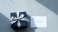 Elegant navy blue gift box with silver ribbon bow beside a thank you card on gray concrete surface with soft shadows.