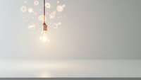Glowing light bulb with innovation icons floating above white surface against gray wall background for creative business concepts.