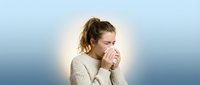 Young woman with light brown hair, Caucasian ethnicity, wearing a cream-colored knit sweater, coughing or sneezing into her hand against a soft blue gradient background. Suitable for healthcare, wellness, and illness-related content.