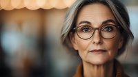 Elegant mature Caucasian businesswoman wearing glasses with confident expression and professional appearance against blurred background with bokeh lights.